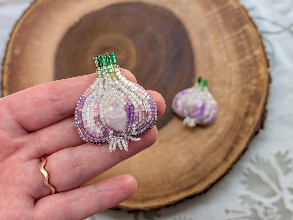 Beaded garlic brooch held in a hand with another brooch on a wooden surface.

