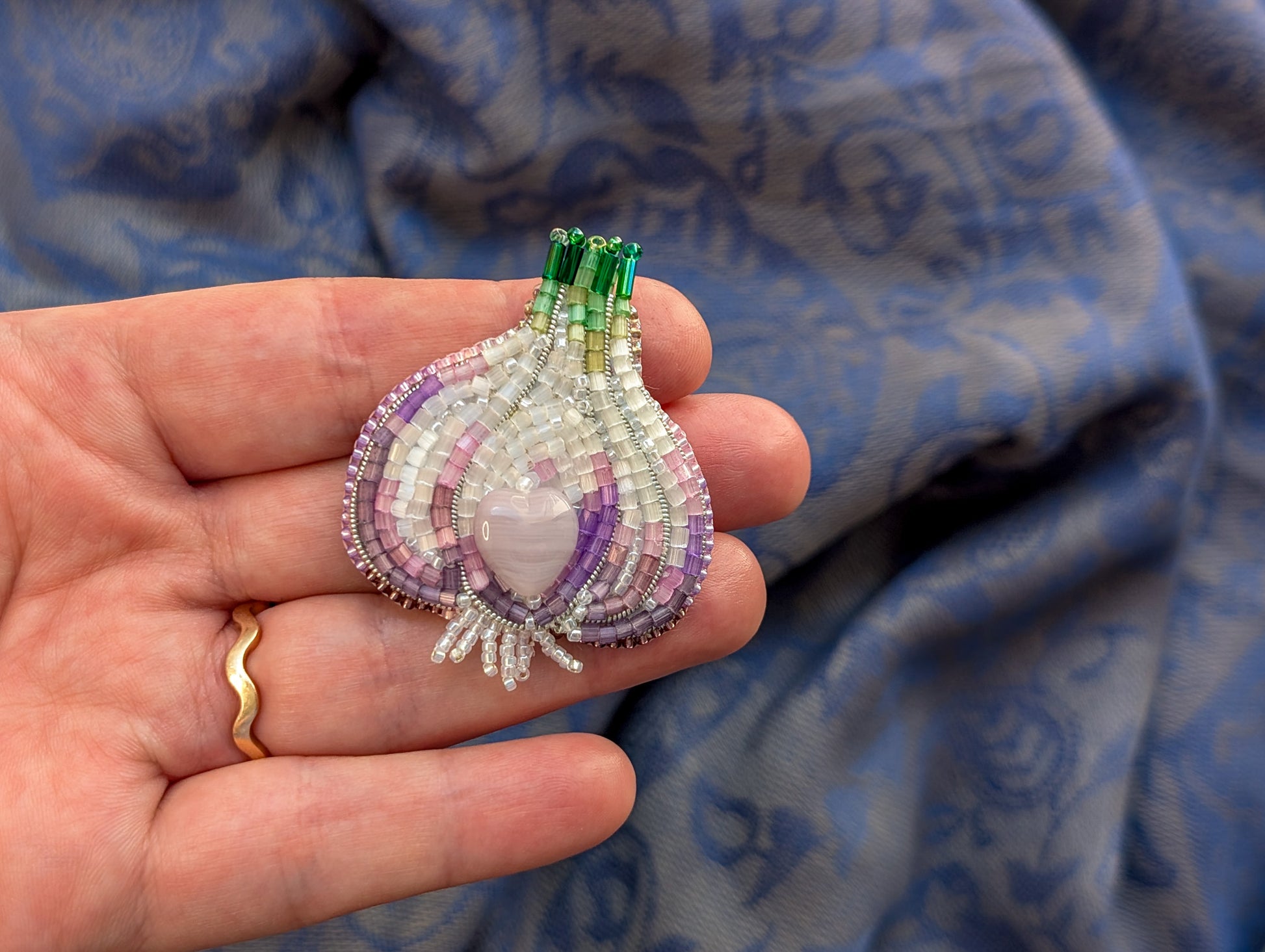 Hand holding a beaded purple and green brooch against a blue fabric background