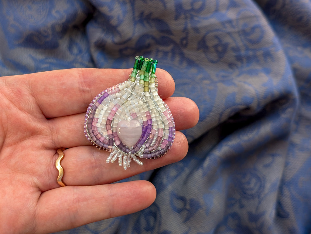 Hand holding a beaded purple and green brooch against a blue fabric background
