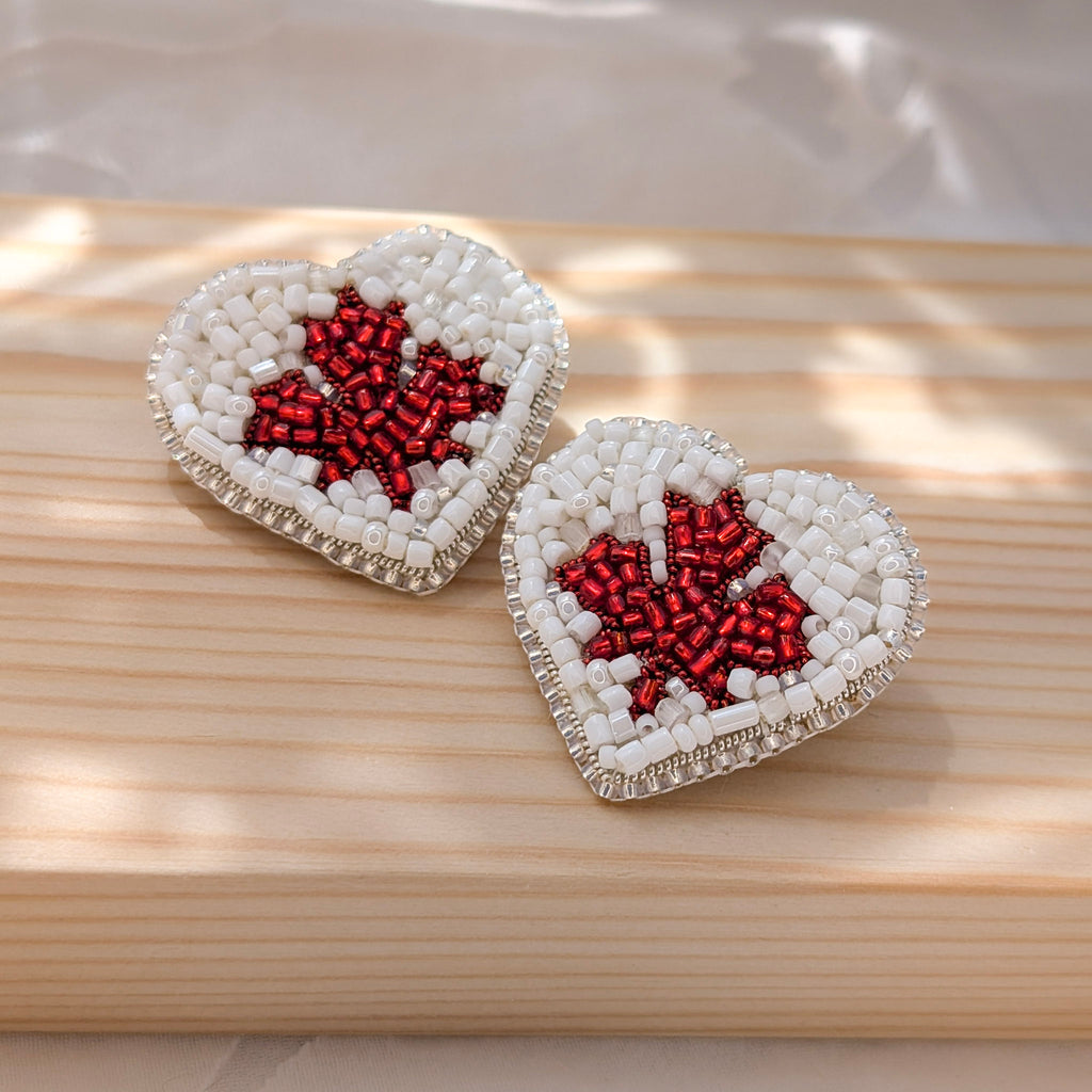 Two heart-shaped pins with a Canadian Maple Leaf design, made of white and red beads on a wooden surface.