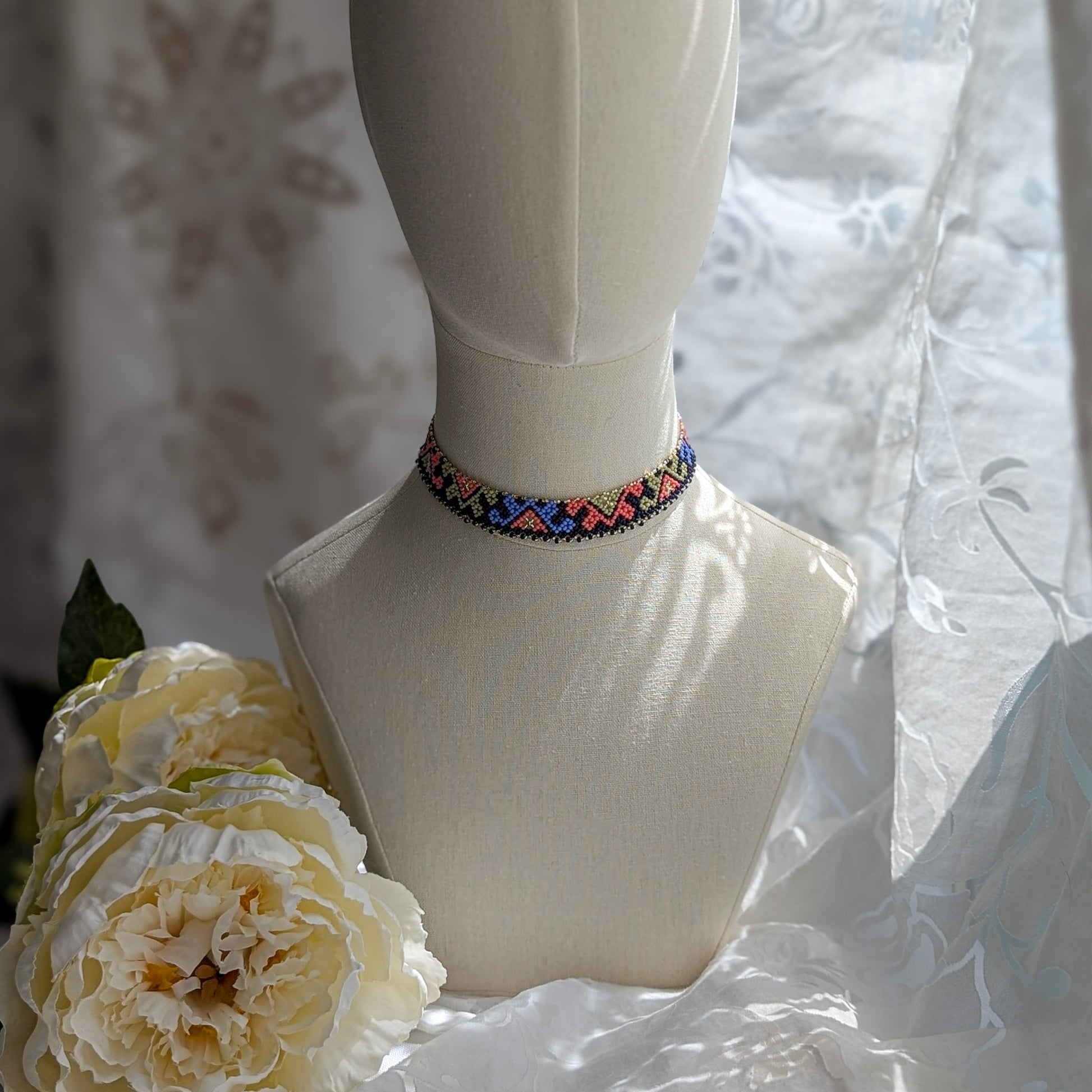 Mannequin with colorful beaded necklace and flowers against a textured white fabric background.